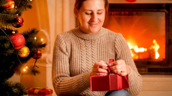 Hermosa mujer sonriente sentada junto a la chimenea ardiendo y atando lazo de cinta en la caja de regalo de Navidad — Foto de Stock