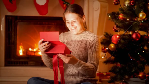 暖炉の横の床に座っている間に輝くクリスマスのギフトボックスの中を見て幸せな笑い若い女性の肖像画 — ストック写真