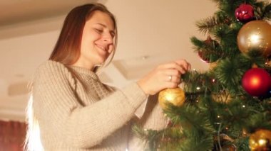Mutlu gülümseyen genç bir kadının oturma odasında takılarla Noel ağacı süslediği 4k yakın çekim videosu.