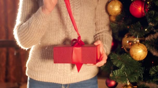 4k βίντεο της νεαρής γυναίκας σε πουλόβερ στέκεται δίπλα στο χριστουγεννιάτικο δέντρο και το άνοιγμα κόκκινο κουτί με δώρο — Αρχείο Βίντεο