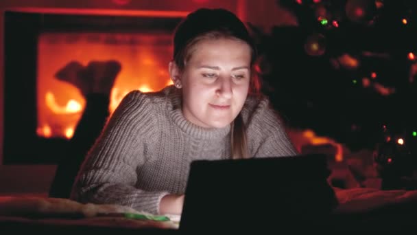 Tablette gülümseyen bir kızın ateş paleti ve parlayan Noel ağacının yanında yattığı 4k video. — Stok video