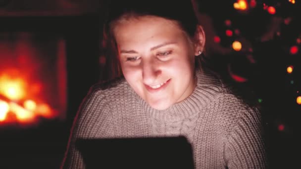 Появилось видео, как улыбающаяся молодая женщина по ночам смотрит видео на планшете в гостиной с горящим камином — стоковое видео