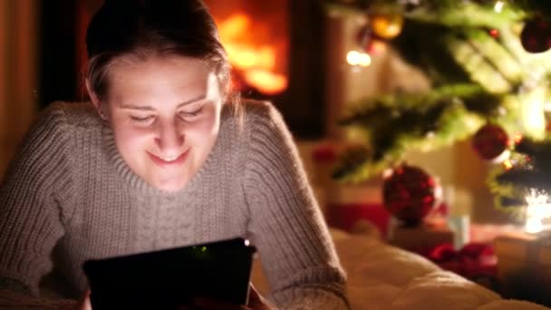 Portrait of happy smiling young woman using laptop while lying next to glowing Christmas tree and fireplace at night — Stock Video