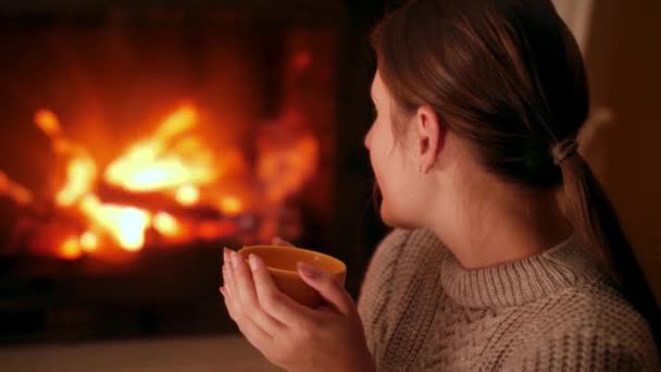 Young woman drinking hot tea and warming at burning fire in fireplace at night in house — Stock Video