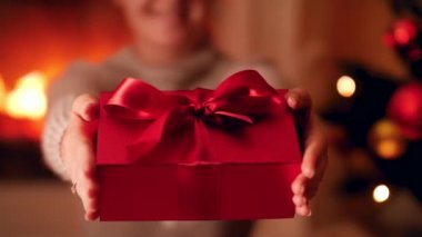 Kapalı 4k video 'da genç bir kadın elinde büyük kurdeleli güzel kırmızı bir hediye kutusu ve Noel ağacı ve şöminenin yanında selam veriyor.