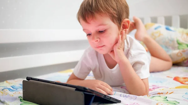 Portrait of cute smiling little boy lying in bed and watching video on digital tablet computer — Stock Photo, Image