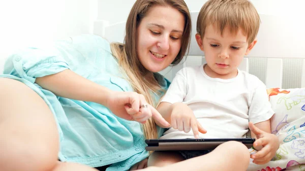 Retrato de menino bonito com sua jovem mãe deitada na cama e navegando na internet no computador tablet digital — Fotografia de Stock