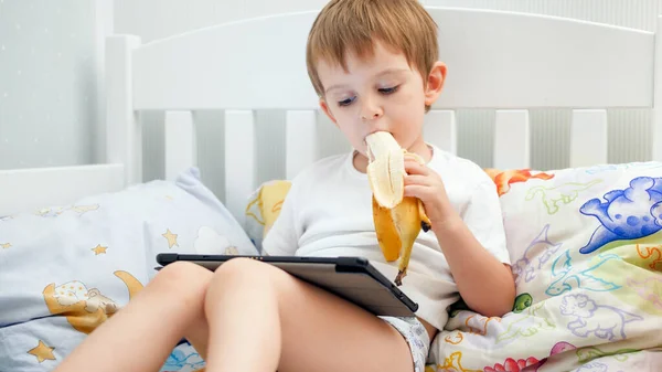 Portrait of cute little boy eating banana and using digital tablet computer before going to sleep — Stock Photo, Image