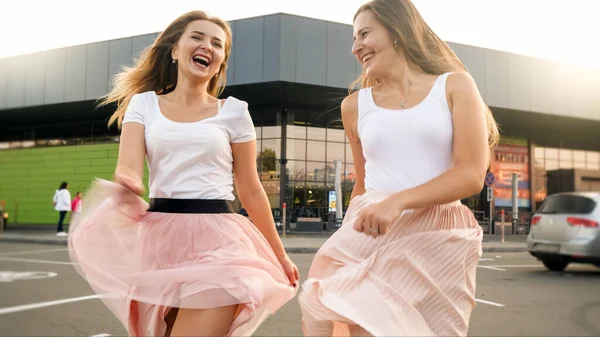 Portrait o two cheerful laughing girls in pink skirt running and jumping on car parking at shopping mall — Stock Photo, Image