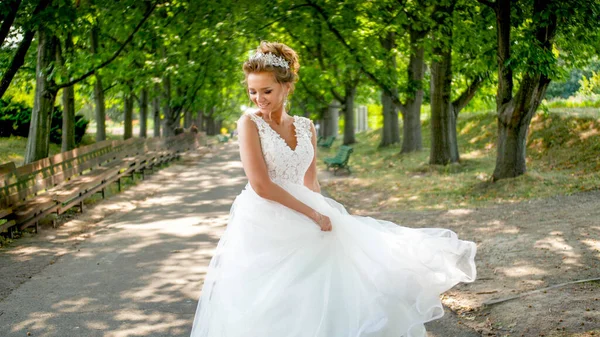 Portret van een vrolijke glimlachende jonge bruid die in het park loopt en zwaait met lange witte bruidsjurk — Stockfoto