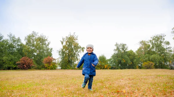 Счастливого улыбающегося мальчика в куртке, бегущего по полю с сухой травой в облачный осенний день — стоковое фото