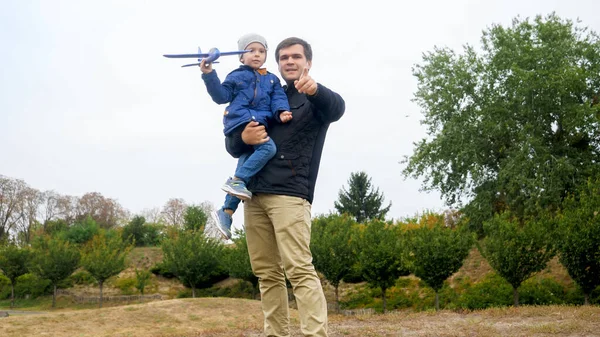 Retrato del joven padre sosteniendo a su pequeño hijo y enseñando a lanzar un avión de juguete al parque — Foto de Stock