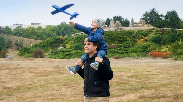 Sonriente niño sentado en los hombros de los padres y lanzando un avión de juguete en el parque —  Fotos de Stock