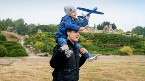 Retrato de criança sorrindo menino sentado no ombro dos pais no parque e lançando avião de brinquedo — Fotografia de Stock