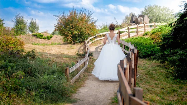 Bela noiva jovem em vestido longo andando no campo ao lado de cerca de madeira longa — Fotografia de Stock