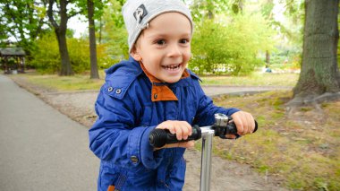 Sonbahar parkında scooter sürerken gülümseyen küçük çocuğun portresi.