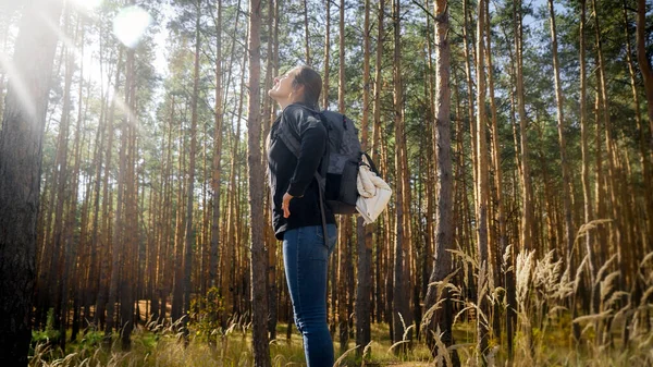 Krásná usměvavá turistka hledící na jasné slunce v borovém lese. Koncept svobody a harmonie s přírodou. — Stock fotografie