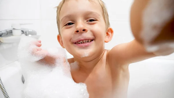 Retrato de niño lindo cubierto de espuma de jabón tomando baño y mirando en la cámara — Foto de Stock