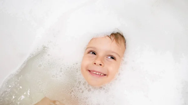 Portrait of cute smiling little boy lying in bath covered with soap foam — Stock Photo, Image