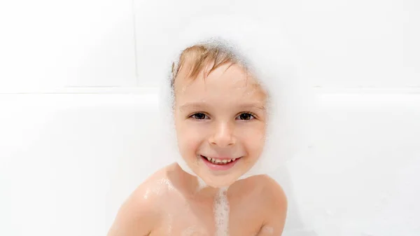 Retrato de lindo niño sonriente con espuma de jabón en la cabeza sentado en el baño y el lavado — Foto de Stock