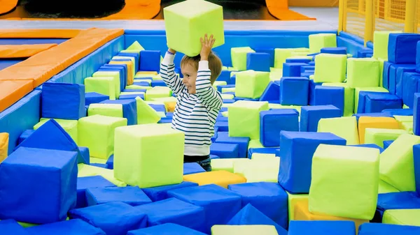 Szczęśliwy uśmiechnięty mały chłopiec bawiący się i rzucający miękkie kostki w dużym dole w pokoju zabaw w centrum handlowym — Zdjęcie stockowe