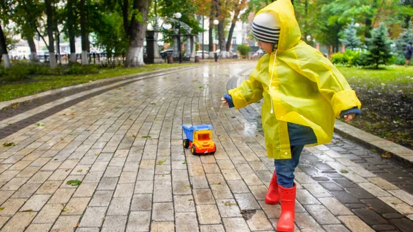 秋雨公园里，穿着黄色雨衣的可爱的小孩开着玩具车跑着 — 图库照片