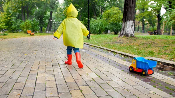 Cute little boy in yellow raincoat running with toy truck under rain at park