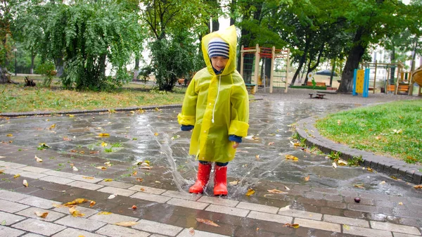 可爱的小男孩穿着雨衣和红色的威灵顿靴在公园的大水坑里跳着 — 图库照片