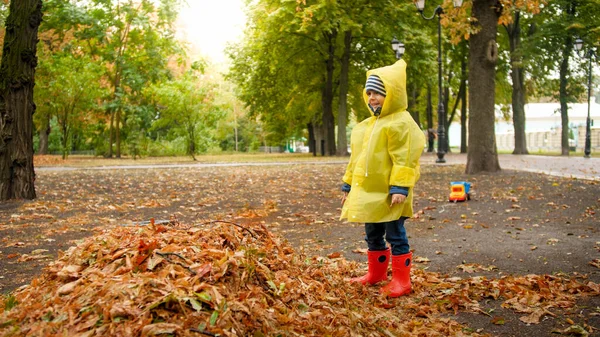 Portrét šťastného smějícího se batolete chlapce ve žlutém plášti a gumových botách si užívá procházky v podzimním parku za deštivého dne — Stock fotografie