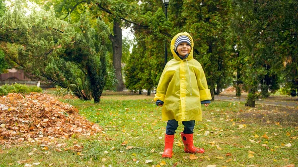 Szczęśliwy uśmiechnięty chłopiec w żółtym płaszczu przeciwdeszczowym i czerwonych gumowych butach po deszczu w parku — Zdjęcie stockowe