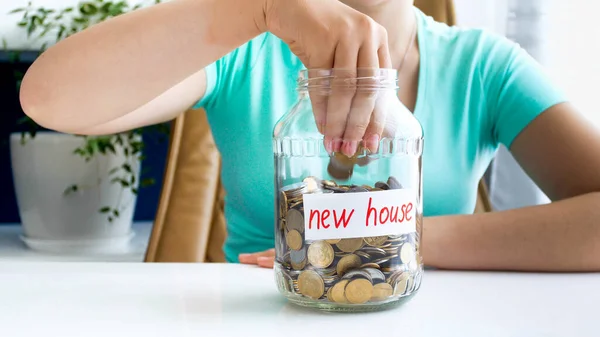 Photo rapprochée de la jeune femme en T-shirt bleu assis à la table blanche sur laquelle se trouve, plein de pièces de monnaie, pot avec l'inscription nouvelle maison — Photo