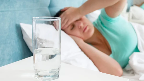 Glass of water on the bedside table against sick woman with migraine lying in bed — Stock Photo, Image