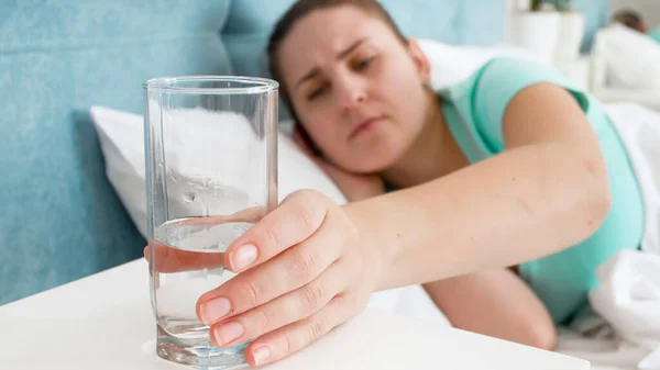 Sick young woman feeling unwell taking glass of water from the bedside table — Stock Photo, Image