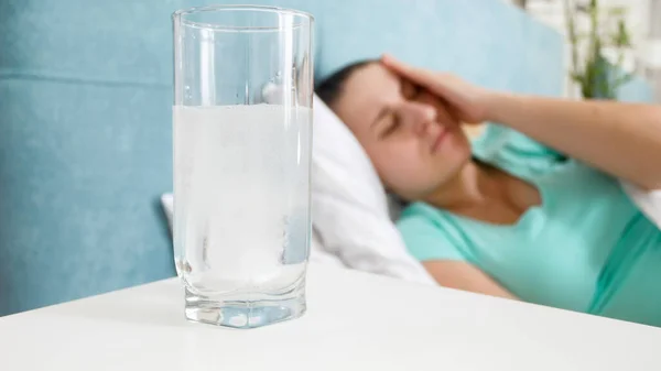 Imagem de close-up de vidro de aspirina contra a mulher deitada na cama sofrendo de dor de cabeça — Fotografia de Stock