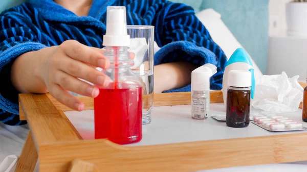 Sick young lying in bed and taking throat spray from medicines tray — Stock Photo, Image