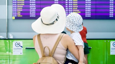 Küçük oğlunu havaalanı terminalinde kucağında tutan ve uçuş çizelgesine bakan genç bir annenin arka plan görüntüsü