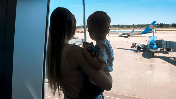 Silhuett av ung kvinna som håller sin son och tittar på flygplan genom fönstret på flygplatsterminalen — Stockfoto