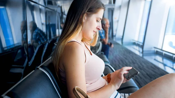 Portrait of beautiful young woman sitting on bench at airport terminal and typing message on smartphone — Stock Photo, Image