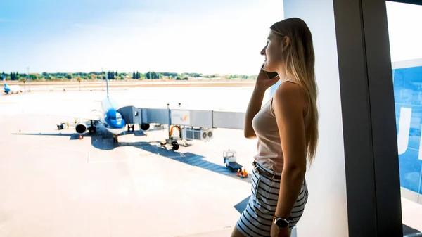 Mooie jonge vrouw staan bij het raam in de terminal van de luchthaven en praten per telefoon — Stockfoto