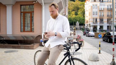 Bisiklete yaslanan ve akıllı telefondan mesaj yazan şık genç adam.