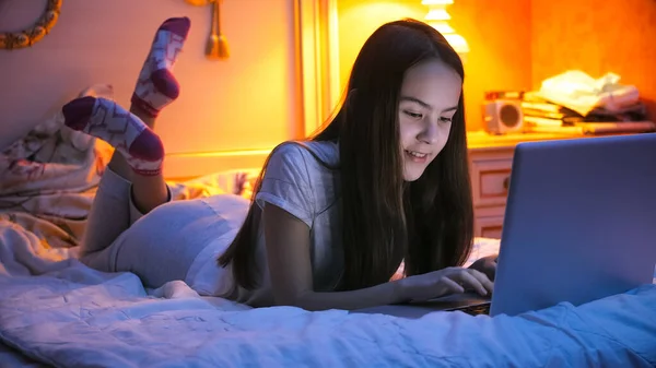 Sorridente ragazza adolescente sdraiata sul letto di notte e chatta sul computer portatile — Foto Stock