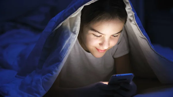 Portrét roztomilé dospívající dívky ležící pod přikrývkou v posteli a pomocí smartphonu — Stock fotografie
