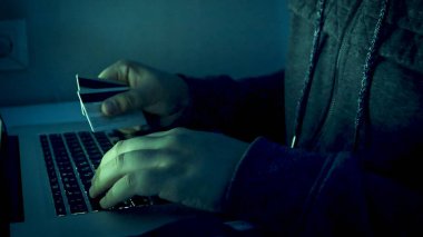 Erkek hackerların ellerinin kredi kartı tutarken ve geceleri dizüstü bilgisayarda yazarken çekilmiş yakın plan görüntüsü.