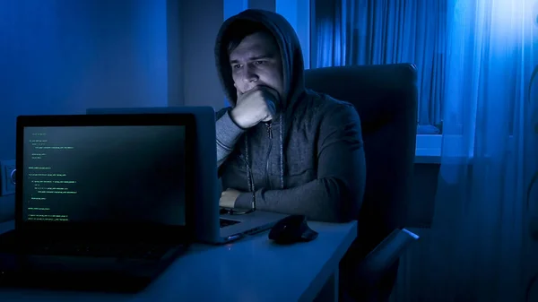 Thoughtful programmer in hood looking on laptop screen while working at night — Stock Photo, Image