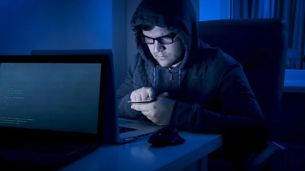 Mężczyzna haker w kapturze trzyma kartę kredytową i pracuje na laptopie w nocy. Cyberprzestępczość i handel elektroniczny — Zdjęcie stockowe