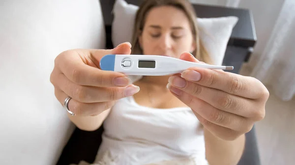 Young sick woman lying on sofa and showing digital thermometer display in camera — Stock Photo, Image