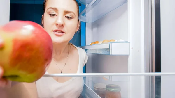 Portrait of hungry woman taking red apple out of fridge on kitchen at night. Concept of dieting and healthy nutrition — Stock Photo, Image