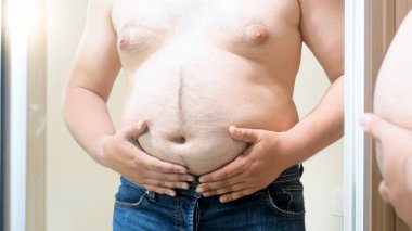 Şişman genç adam büyük aynada duruyor ve koca göbeğini tutuyor. Erkek aşırı kilolu, kilo kaybı ve diyet kavramı
