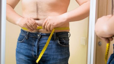 Kıllı göbekli genç bir adam, karnını mezurayla ölçüyor. Erkek aşırı kilolu, kilo kaybı ve diyet kavramı