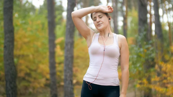 Portrait of tired young woman touching forehead after jogging in forest or park. — Stock Photo, Image
